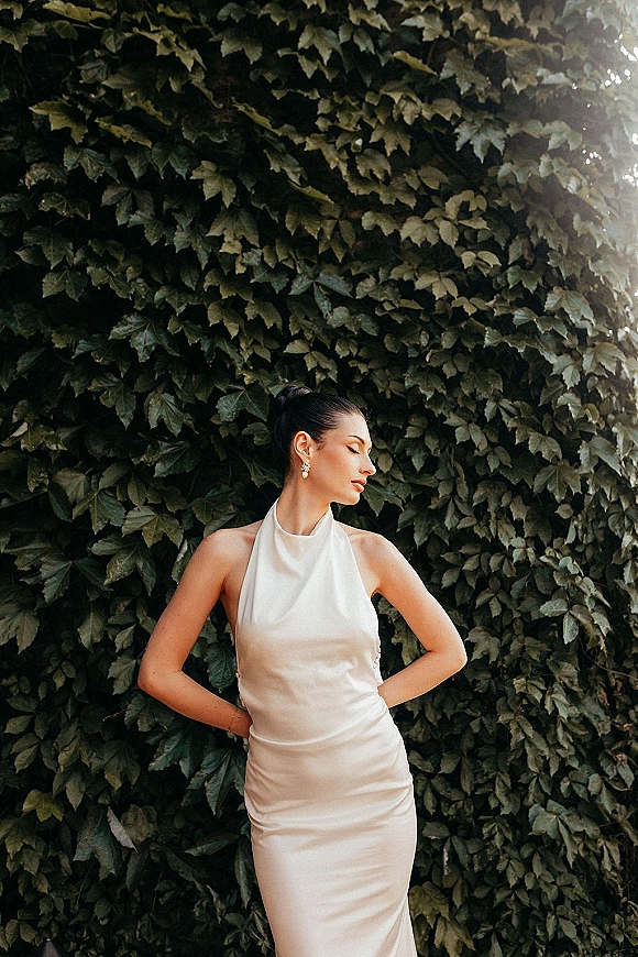 Bridal portrait of a bride in a halter wedding dress with drop earrings and updo, hands on hips beside an ivy wall in natural light