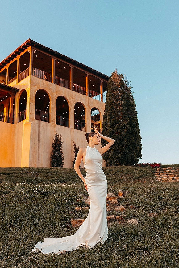 Bridal portrait in a halter wedding dress with a long train, hair in a bun and earrings, standing on villa stone steps at sunset