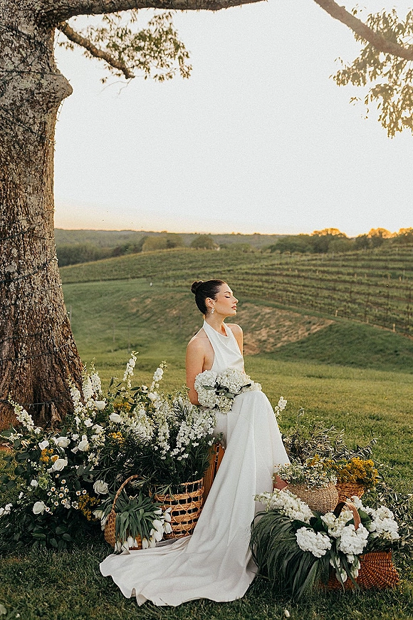 Bridal portrait of a bride in a halter wedding dress holding a white bridal bouquet, seated under a large tree at sunset vineyard hills