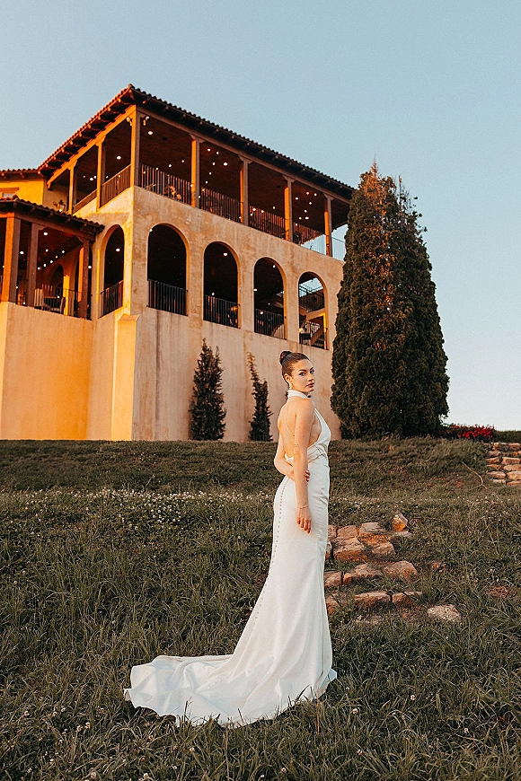 Bridal portrait of a bride in a halter wedding dress with open back and train, looking over her shoulder on stone steps at a stucco villa balcony with string lights