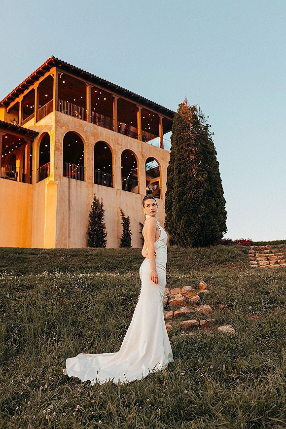 Bridal portrait of a bride in a simple satin wedding dress with a long train, looking over her shoulder on stone steps by an arched villa balcony