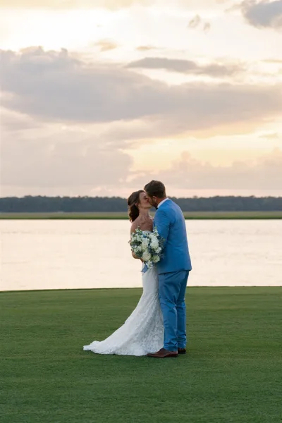 Wedding kiss portrait of bride and groom kissing at sunset by a lake, bride holding a white rose bouquet, groom in a blue suit