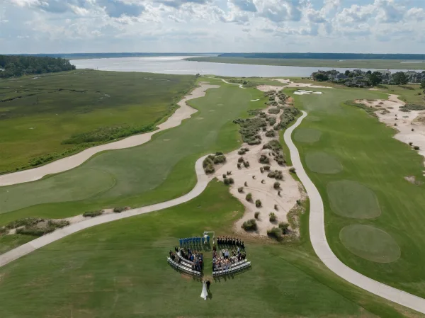 Outdoor wedding ceremony, aerial wedding ceremony view with semicircle chairs and a simple arch on a coastal golf course by marsh and water
