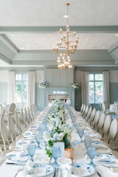 Reception tablescape on a long banquet table setup with blue goblets, blue-and-white plates, and floral runner beneath a chandelier by tall windows