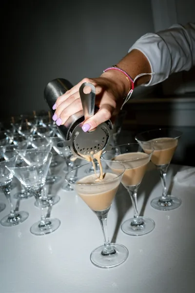 Wedding cocktail service as a hand with lilac manicure strains creamy cocktails into martini glasses on a white tablecloth in a dim room