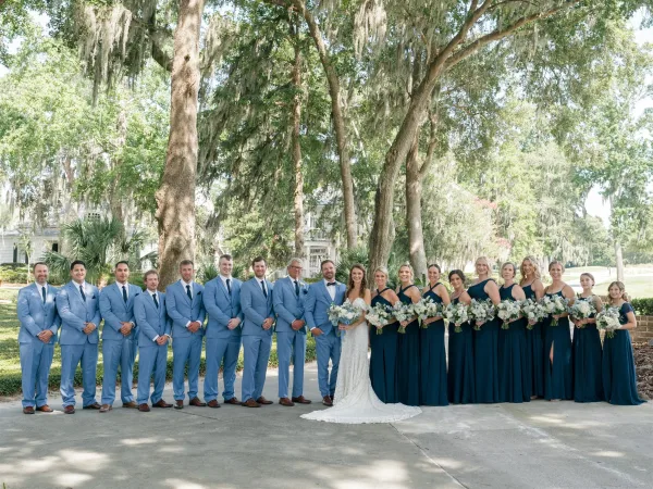 Wedding party portrait with bride and groom flanked by bridesmaids in navy dresses and groomsmen in light blue suits under oak trees with Spanish moss
