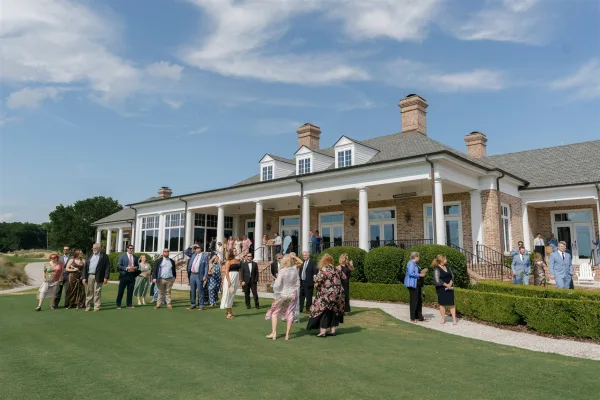 Wedding guests mingle at cocktail hour with drinks and sunglasses on a lawn outside a brick clubhouse with white columns under blue sky