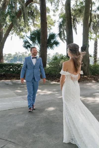 First look moment as bride in an off the shoulder lace open-back gown walks to groom in bow tie on a tree-lined path by the lake