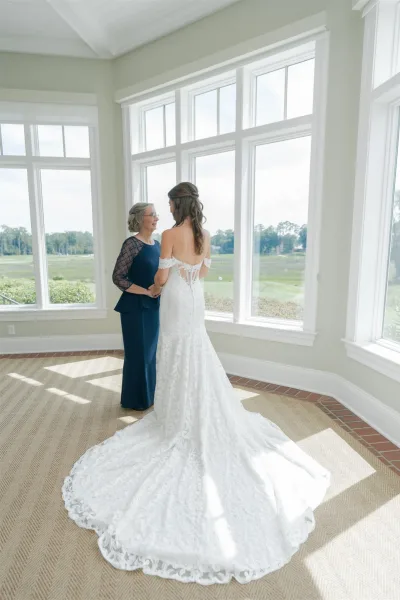 Bride with mother sharing a mother of the bride first look, holding hands by large windows in natural light, lace off-shoulder gown train