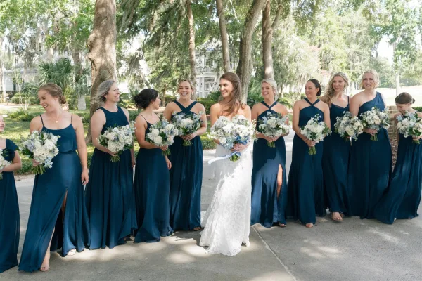 Bridal party portrait with bride with bridesmaids holding white and blue bouquets, standing on a paved walkway under oak trees and moss