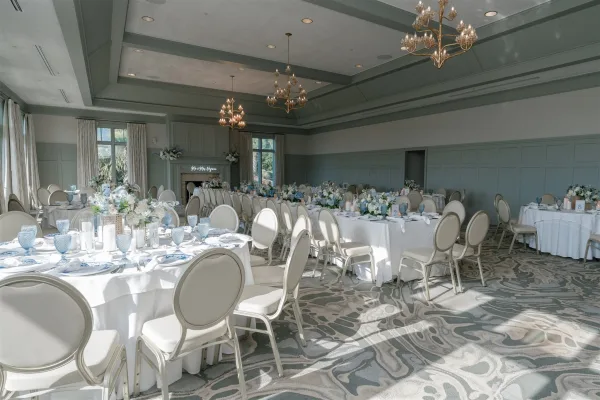Reception tablescape with round wedding reception tables in white linens, blue goblets, floral centerpieces and candles under chandeliers in a paneled banquet hall