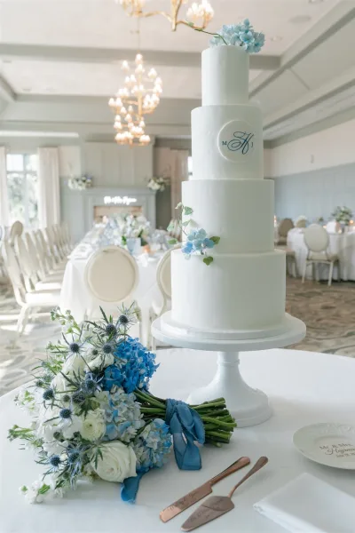 Wedding cake four tier wedding cake in white buttercream with blue sugar flowers, greenery, and monogram topper on a stand in chandelier-lit reception room