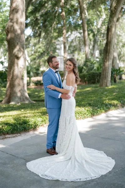 Couple portrait of bride in a lace wedding dress and groom in a blue suit embracing on a sunlit, tree-lined garden path