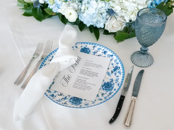 Reception tablescape with blue and white wedding tablescape china, white napkin and menu card on plate, blue goblet, and hydrangea rose centerpiece