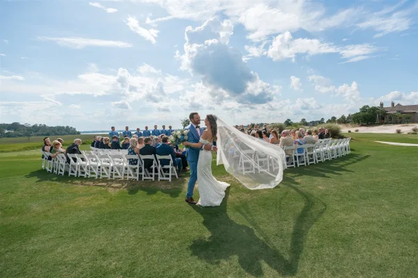 Ceremony moment as bride and groom embrace, her long veil blowing in the wind while guests sit on white chairs by the water under a dramatic sky