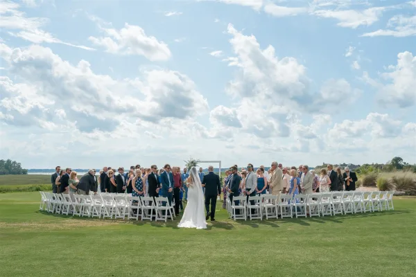 Outdoor wedding ceremony with bride walking down the aisle in a long veil as guests stand by white folding chairs near a waterfront marsh view