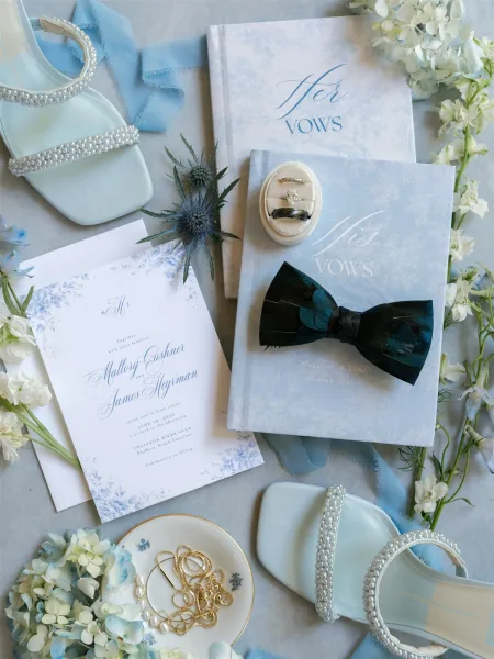 Wedding flat lay with wedding invitation suite, vow books, pearl shoes, rings, bow tie, jewelry, and blue hydrangea on light fabric surface