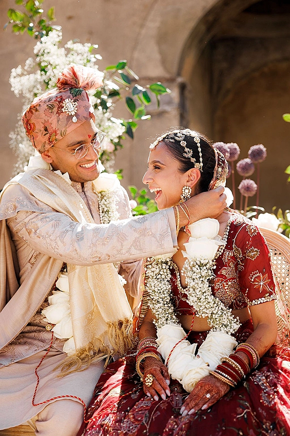 Wedding couple portrait of a Hindu wedding couple in white floral garlands, laughing beneath a stone archway in a lush outdoor courtyard
