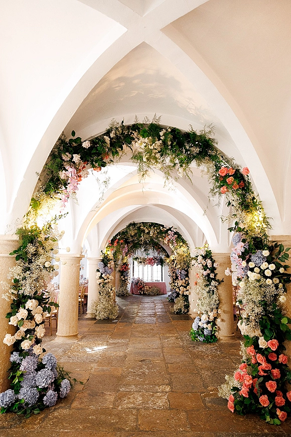 Floral ceremony arch with garden rose blooms and hydrangeas, framing a stone corridor with vaulted ceiling and soft window light