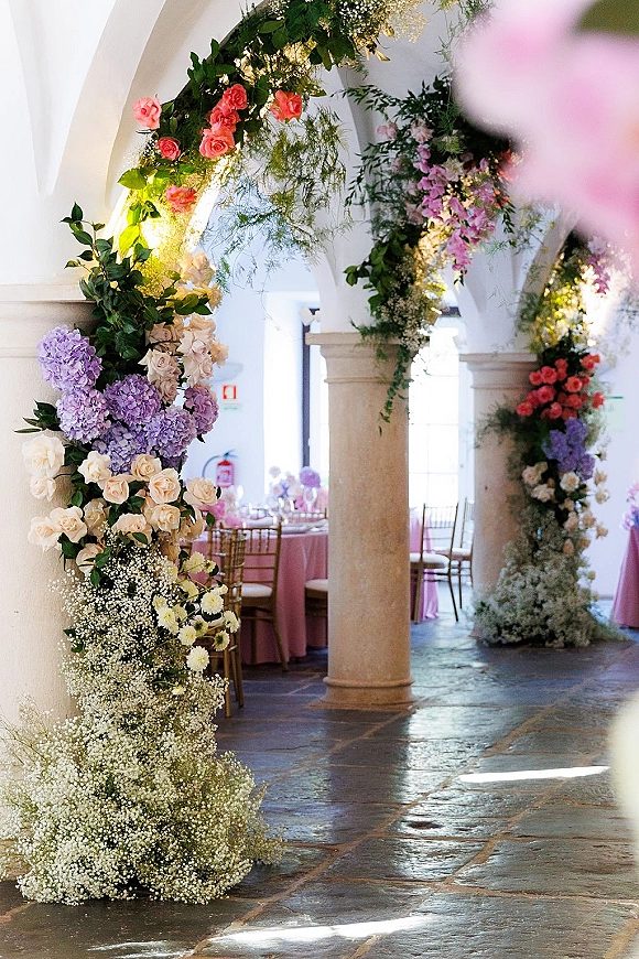 Wedding floral arch with roses and hydrangeas draped in greenery over white arches and stone columns, with pink tables in back
