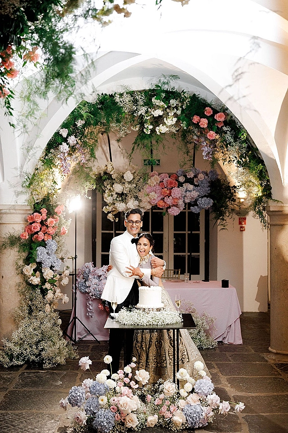 Cake cutting moment as bride and groom slice a white wedding cake at a floral-arched cake table with champagne flutes in a courtyard patio
