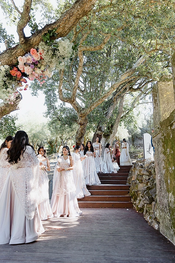 Bridesmaid procession in champagne dresses carrying blush bouquets down wooden stairs lined with rose petals beneath oak trees