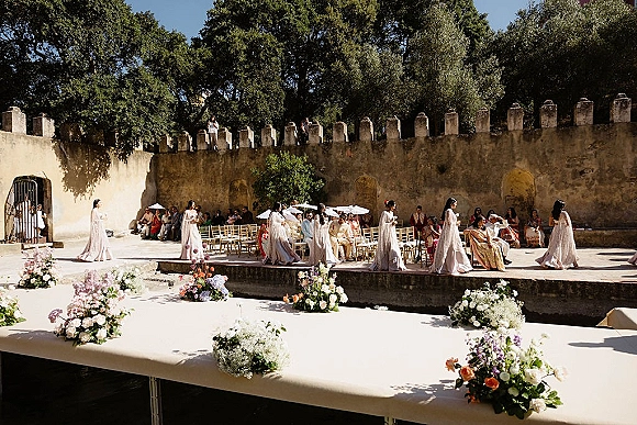 Outdoor ceremony setup with courtyard wedding ceremony seating, aisle flowers and parasols as bridesmaids walk past a stone wall backdrop