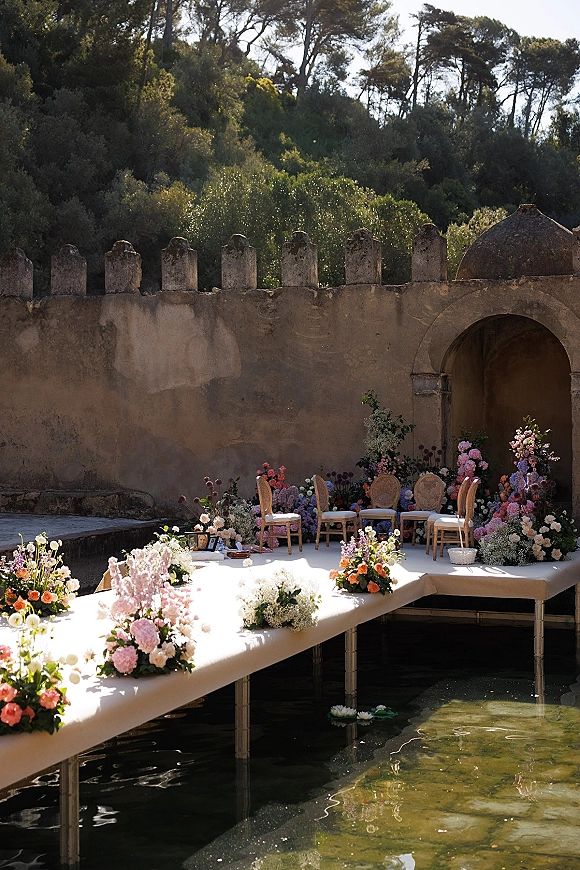 Ceremony setup with a poolside wedding ceremony, featuring a white platform aisle over water with floating floral decor by stone walls