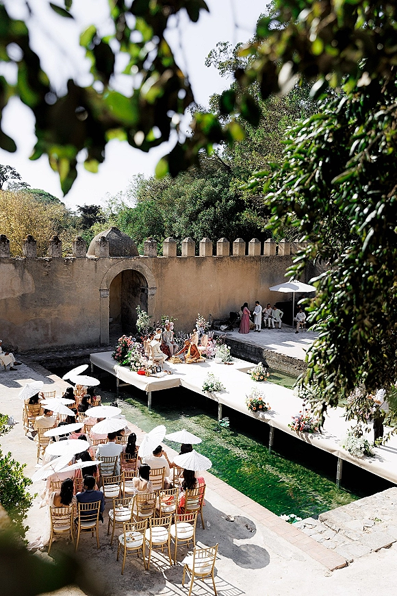 Outdoor wedding ceremony with a white aisle platform over a canal, floral arrangements and parasols, gold chairs in a stone courtyard with arched doorway