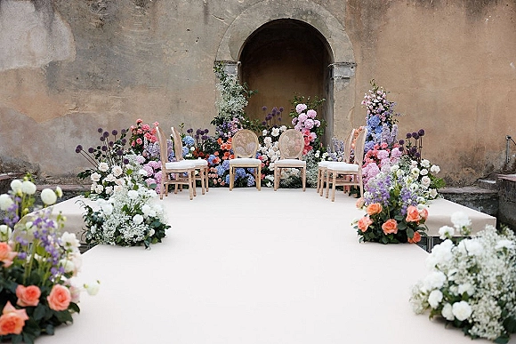 Ceremony setup with a white wedding aisle runner, flower-lined ground clusters and cane-back chairs before a stucco archway doorway.