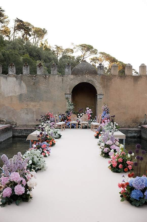 Ceremony aisle design with floral lined wedding aisle and a white aisle runner leading to an arched doorway by a reflecting pool
