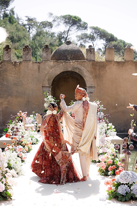 Wedding recessional as bride and groom walk away hand in hand, flower petals on a white aisle runner in an outdoor courtyard stone archway