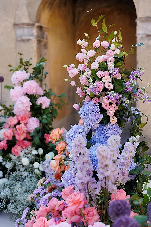 Wedding floral installation with pink roses and blue hydrangeas nestled in greenery at the base of a stone archway against stucco wall