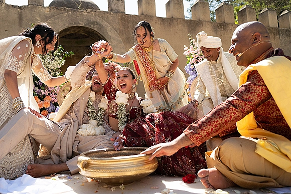Wedding ceremony moment at an Indian wedding ceremony as bride and groom laugh under rose petals in a sunlit courtyard by a stone wall