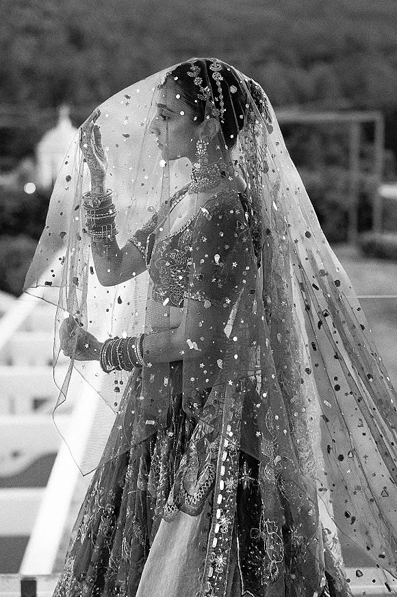 Bridal portrait in black and white of a bride in side profile under a veil, wearing an embellished lehenga on outdoor steps with a blurred garden backdrop