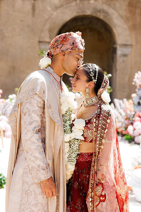 Wedding couple portrait of groom kissing bride’s forehead beneath a stone archway, both in ornate South Asian attire with floral garlands
