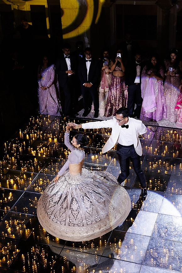 First dance on a wedding dance floor as groom in white dinner jacket spins bride in lehenga under string lights on a mirrored floor