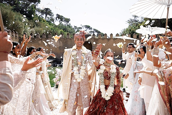 Wedding recessional during a hindu wedding exit as the newlyweds raise hands, wearing garlands while guests toss petals in a courtyard