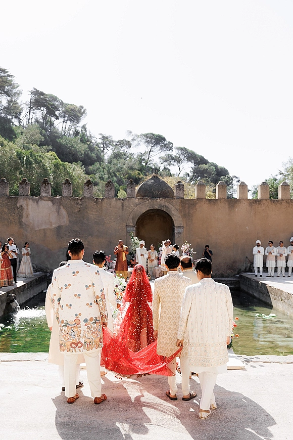 Wedding ceremony moment at an indian wedding ceremony as bride walks with red veil past reflecting pool in a stone courtyard with arches