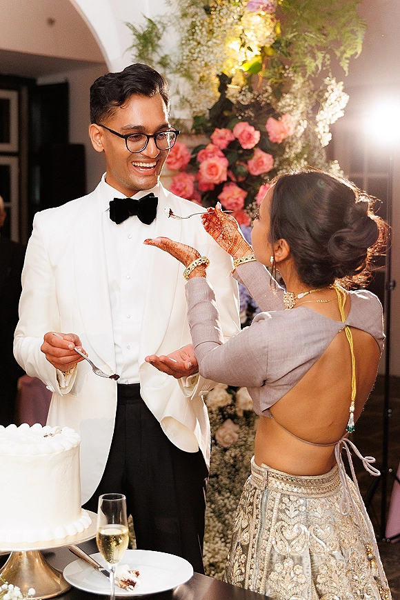Wedding cake cutting as the bride feeds groom cake, hennaed hands holding a fork beside champagne flute and rose greenery under warm arches
