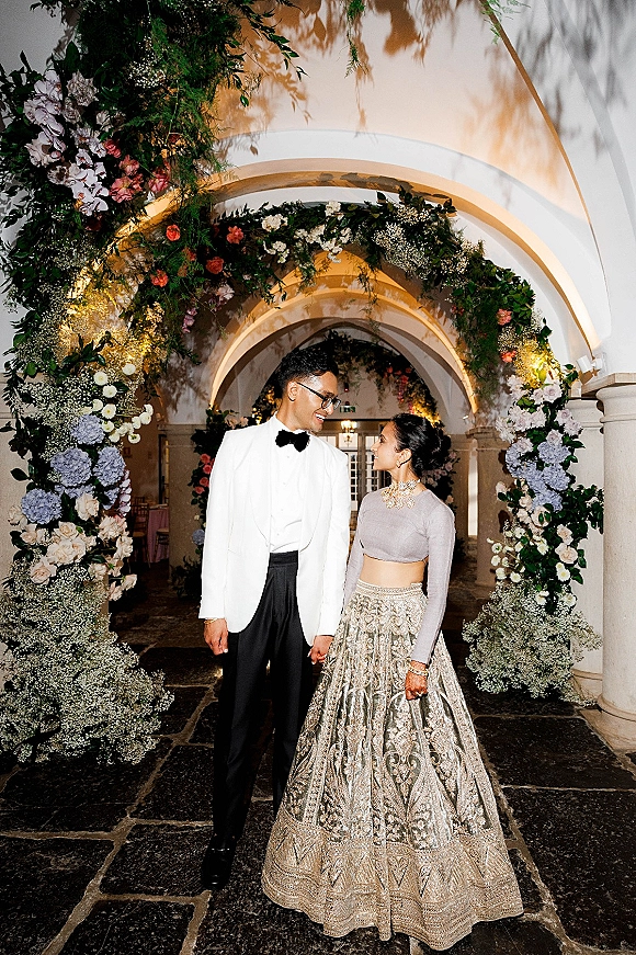 Couple portrait of bride and groom holding hands beneath a floral arch of hydrangeas and roses in an arched indoor hallway corridor.