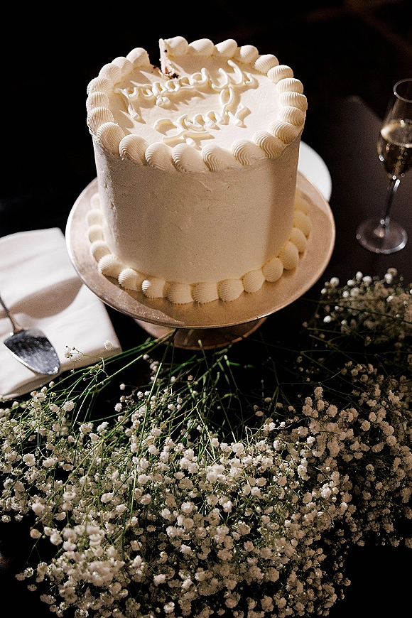 Wedding cake with piped buttercream border on a stand, featuring an inscription, cake knife, baby's breath, greenery, and champagne flute on dark tabletop