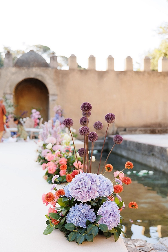 Ceremony aisle florals with hydrangeas, allium and orange blooms line a white runner between chairs in a stone courtyard by water.