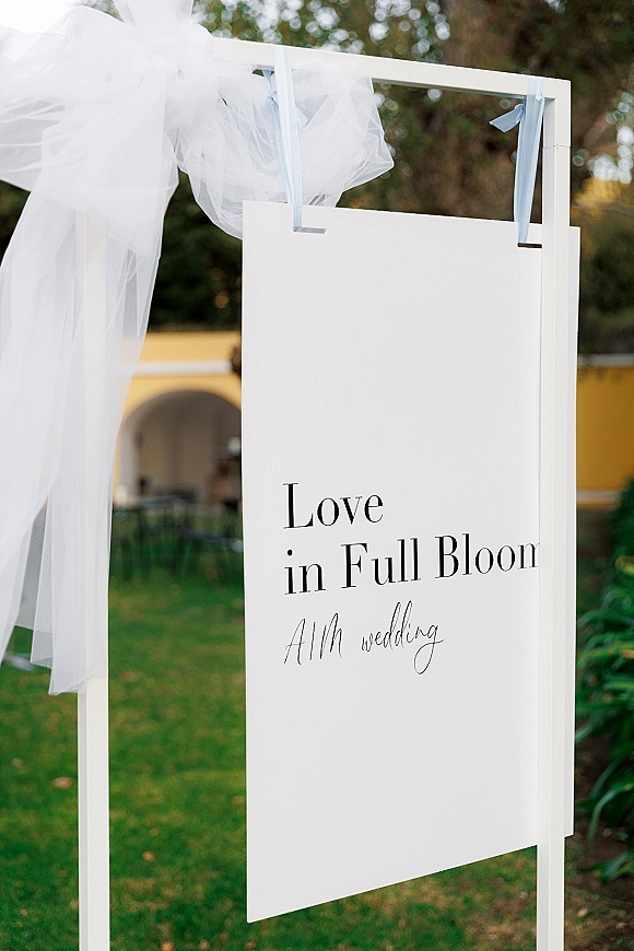 Wedding welcome sign with black typography on a white board, hanging from a stand with blue ribbon, set on a garden courtyard lawn