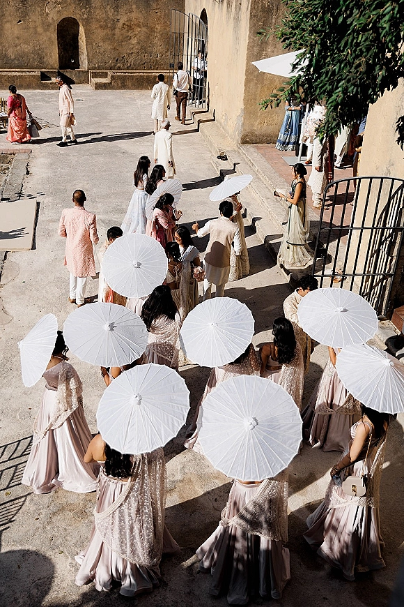 Wedding procession with a bridal party entrance under white parasols, walking through a stone courtyard toward an arched doorway in shade
