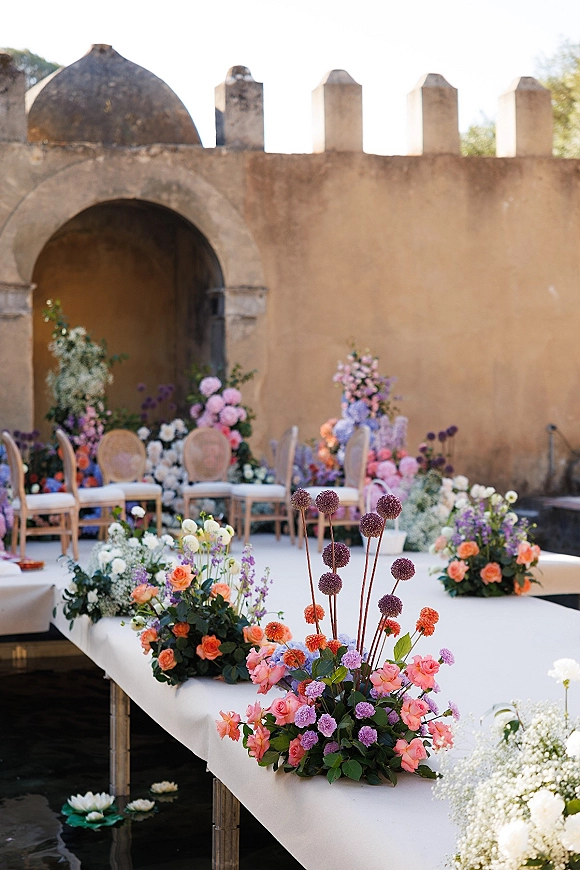 Ceremony aisle decor with a white aisle runner and ground floral arrangements of roses, hydrangea, and greenery in a courtyard with arched doorway