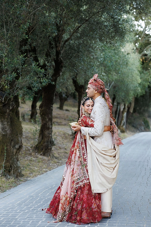 Couple portrait of a south asian wedding couple embracing, bride in a red lehenga and groom in embroidered sherwani and turban on a tree-lined path
