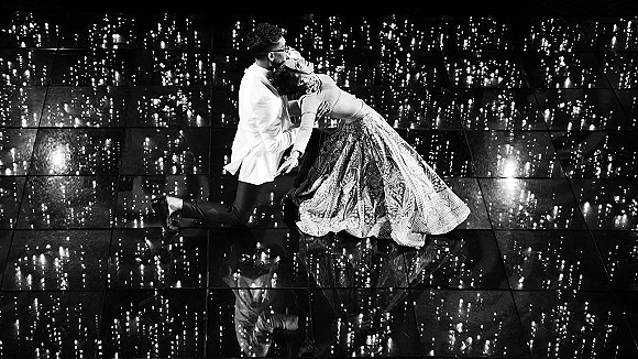 Couple portrait in a wedding dance pose with the groom kneeling as the bride leans back, lehenga skirt reflected on a mirror floor under twinkle lights