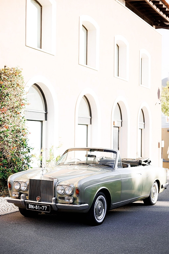 Wedding getaway car, a silver vintage convertible with chrome grille parked by a white stucco building with arched windows and greenery