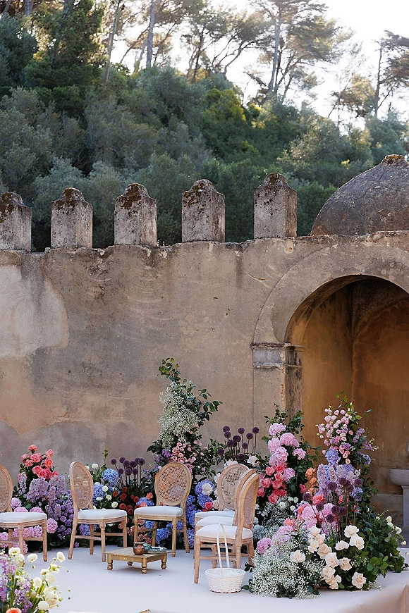 Ceremony backdrop with ground florals of roses, hydrangeas and greenery framing cane chairs, with a stone arch and stucco wall behind